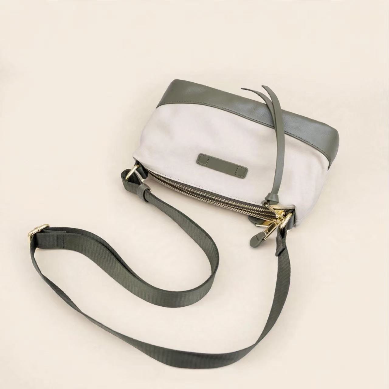 A small crossbody bag with a white body, dark gray top panel and strap, and gold-tone zipper hardware, photographed on a plain light beige background.